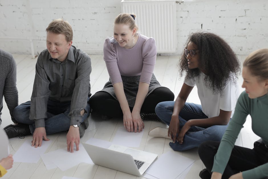 A diverse group of adults engaged in a discussion, sitting on the floor with papers and a laptop